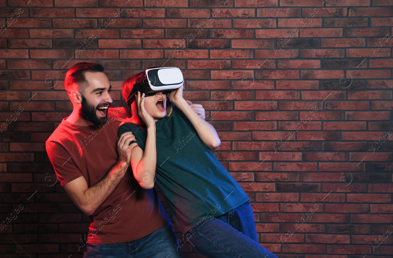 Photo of Emotional woman playing video games with VR headset and young man near brick wall. Space for text