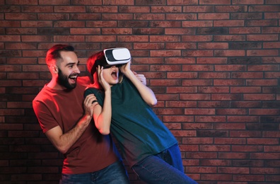 Emotional woman playing video games with VR headset and young man near brick wall. Space for text