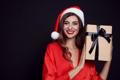Photo of Happy woman in Santa hat with gift box on black background, space for text. Christmas party