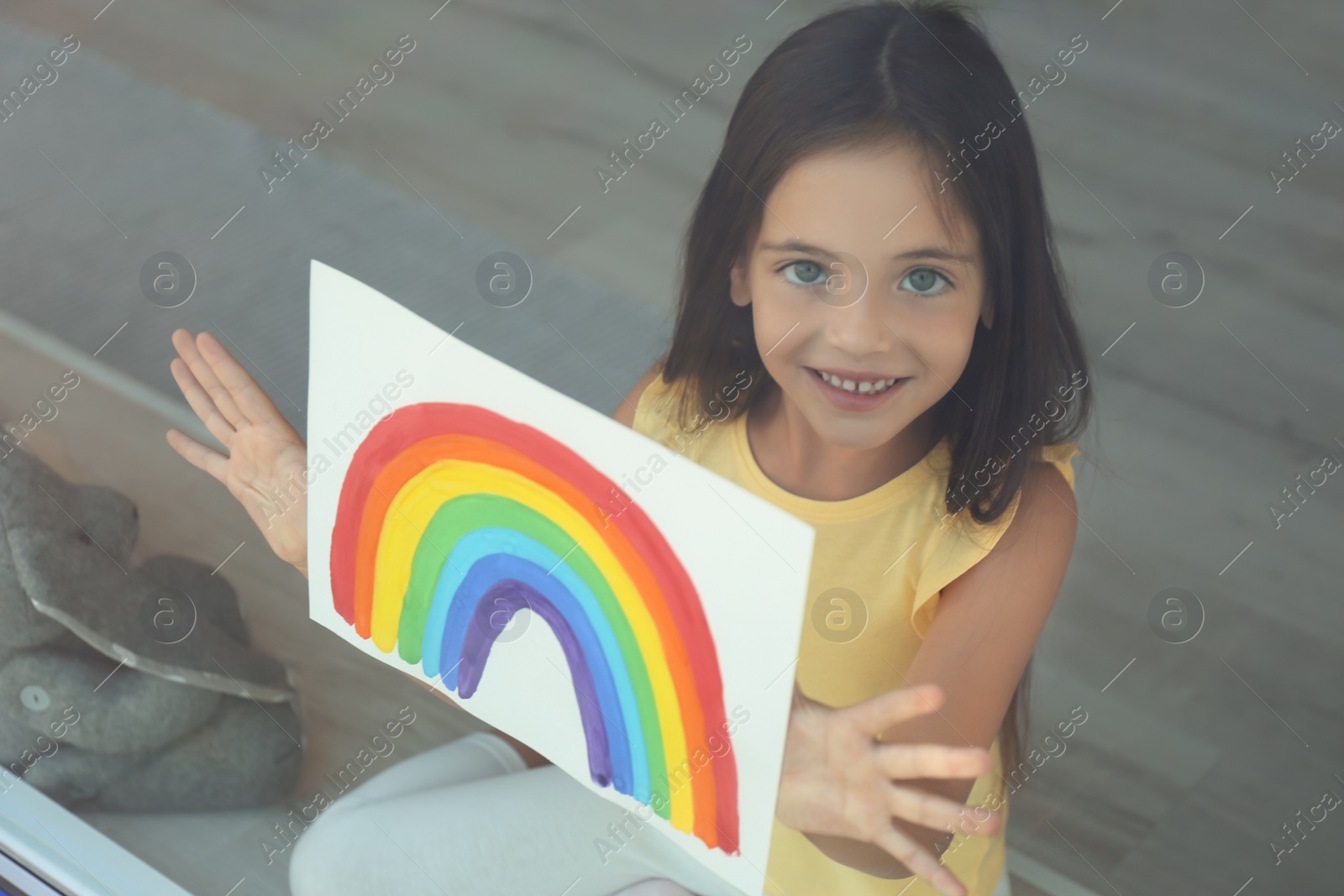 Photo of Little girl with picture of rainbow near window, view from outdoors. Stay at home concept