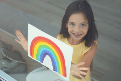 Little girl with picture of rainbow near window, view from outdoors. Stay at home concept