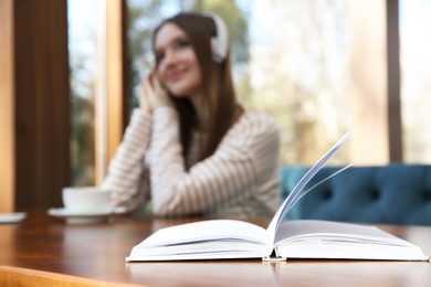 Photo of Book on table in cafe and woman with headphones on background. Audiobook concept