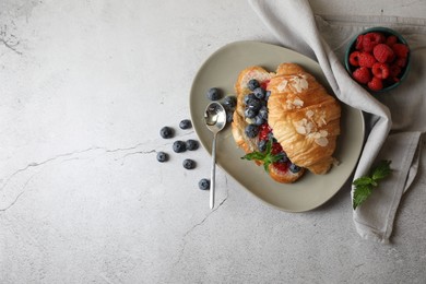 Delicious croissant with berries, almond flakes and spoon on light grey table, flat lay. Space for text