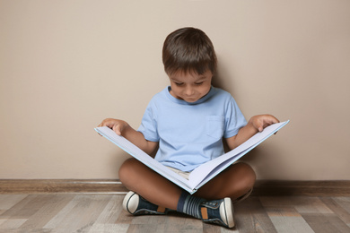 Photo of Cute little boy reading book on floor near beige wall