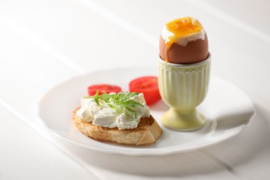 Fresh soft boiled egg in cup and sandwich served on white wooden table. Space for text