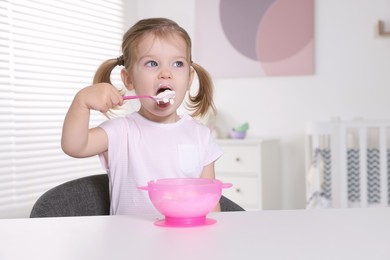 Cute little child eating tasty yogurt from plastic bowl with spoon at white table indoors