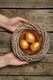 Woman holding nest with shiny golden eggs at wooden table, top view