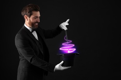 Magician showing trick with wand and top hat on dark background. Fantastic light coming out of hat