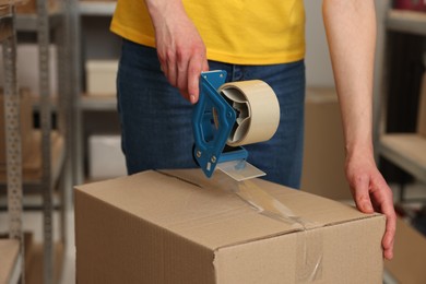 Photo of Post office worker packing box with adhesive tape indoors, closeup