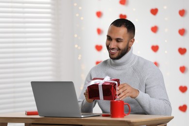 Valentine's day celebration in long distance relationship. Man holding gift box while having video chat with his girlfriend via laptop indoors