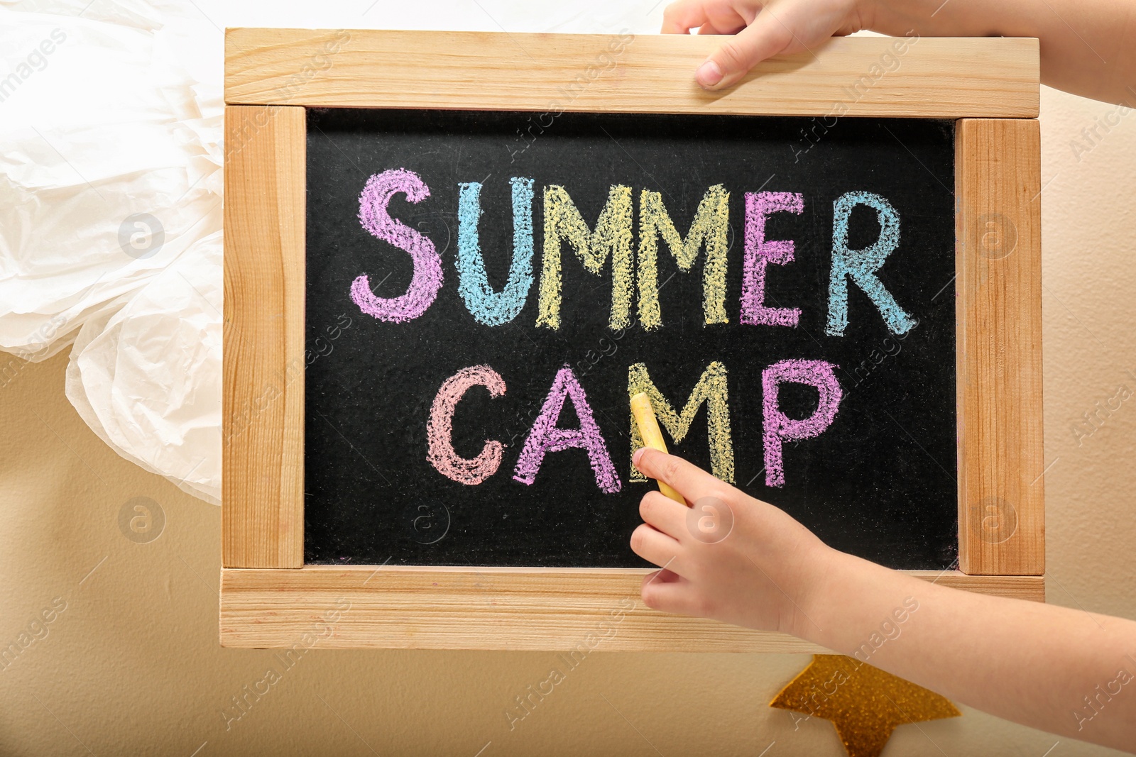 Photo of Child writing SUMMER CAMP with colorful chalk on blackboard, closeup