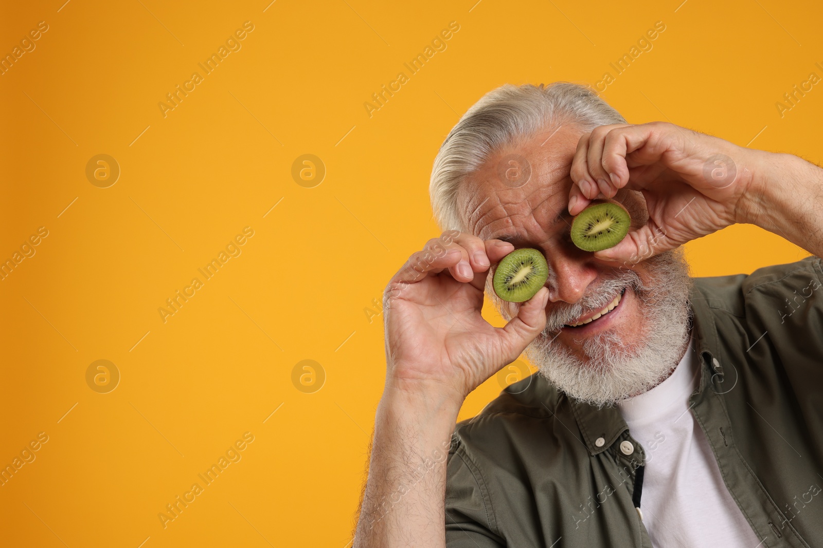 Photo of Senior man covering eyes with halves of kiwi on orange background, space for text