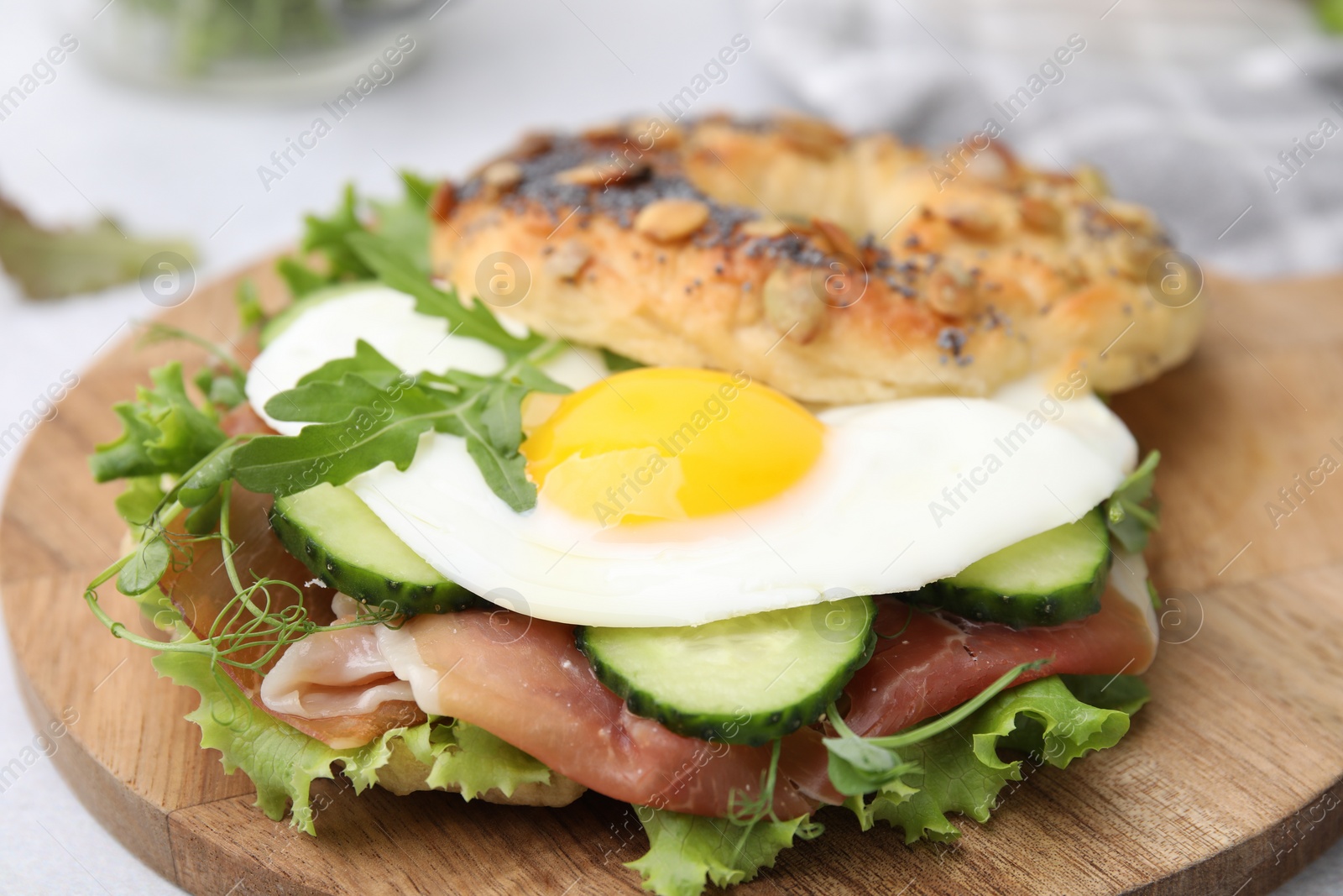 Photo of Tasty bagel with cured ham, egg, cucumber and salad mix on wooden board, closeup