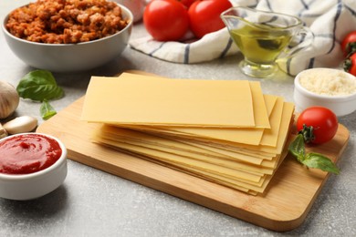 Cooking lasagna. Wooden board with pasta sheets and other products on grey table, closeup