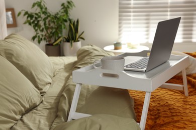 Photo of White tray with modern laptop and cup of aromatic drink on bed indoors, space for text