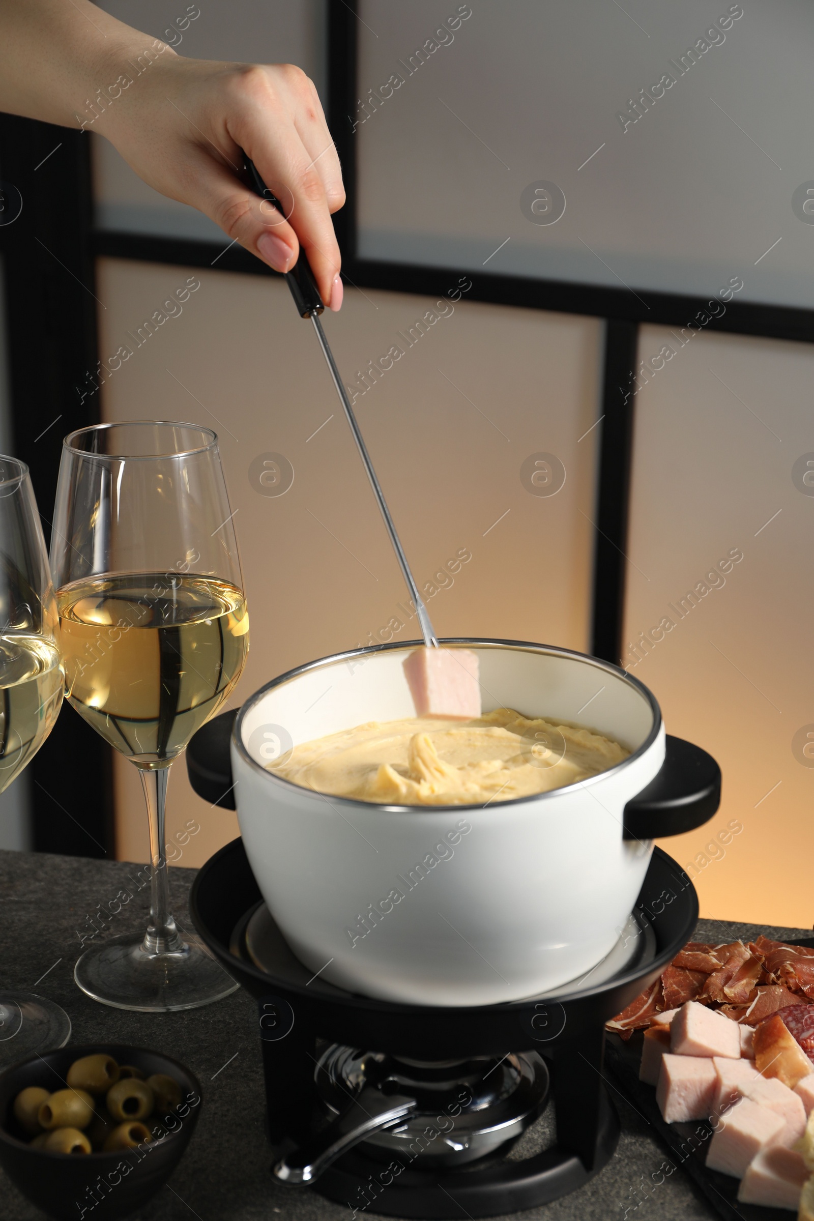 Photo of Woman dipping piece of ham into fondue pot with melted cheese at grey table with wine and snacks, closeup