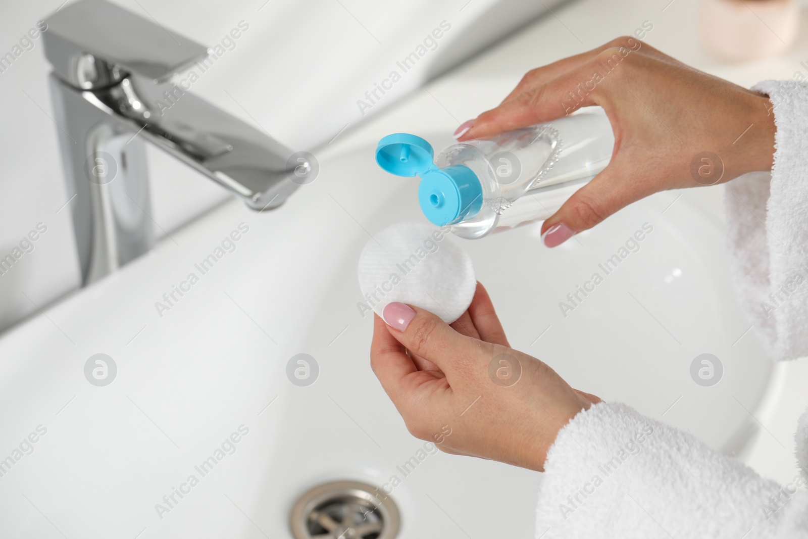 Photo of Woman pouring makeup remover from bottle onto cotton pad near sink indoors, closeup