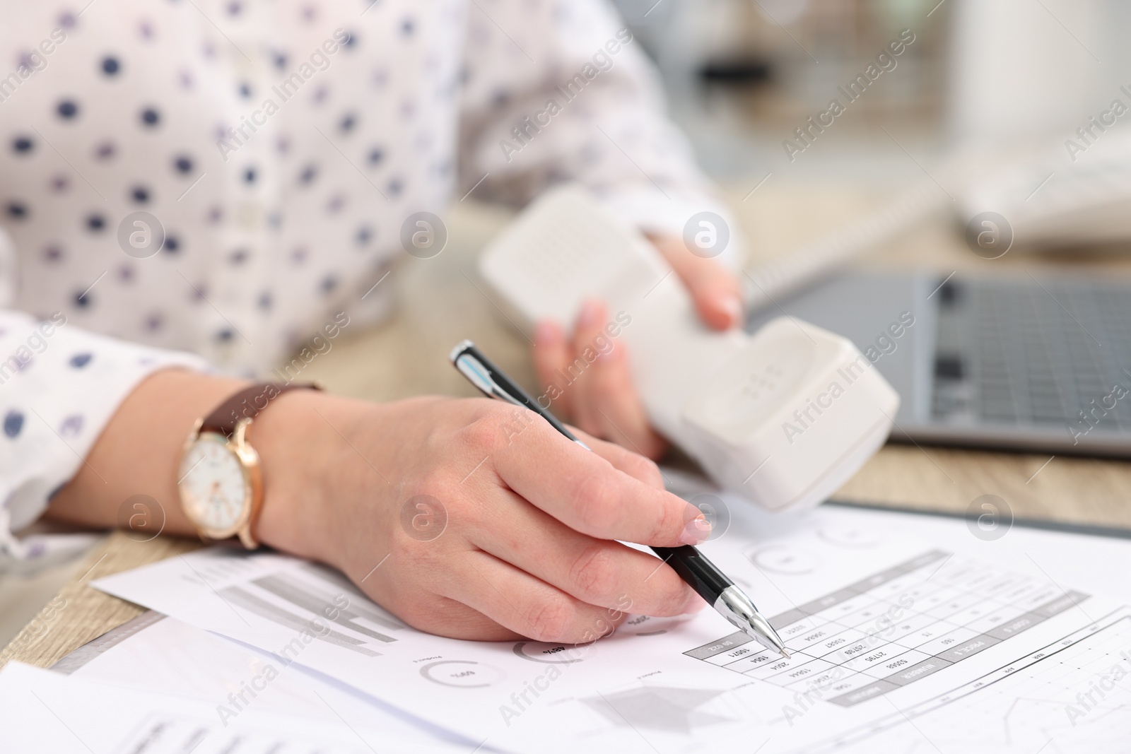 Photo of Secretary with telephone handset doing paperwork at table in office, closeup