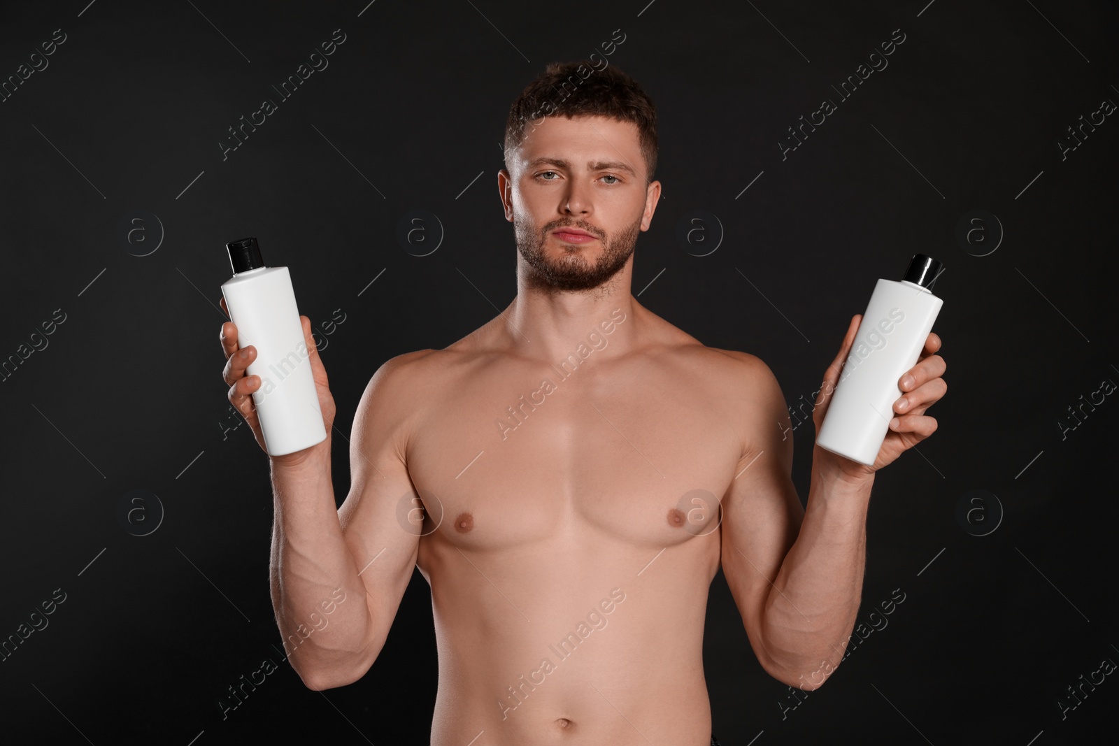 Photo of Shirtless young man holding bottles of shampoo on black background