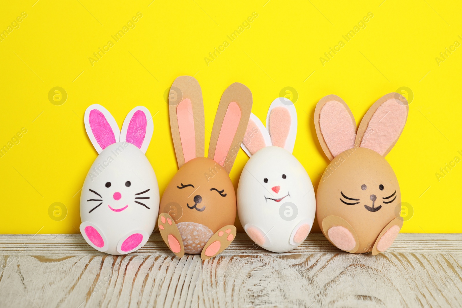 Photo of Eggs as cute bunnies on white wooden table against yellow background. Easter celebration