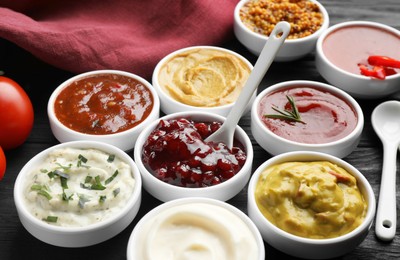 Photo of Different tasty sauces in bowls and spoons on black wooden table, closeup