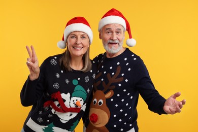 Happy senior couple in Christmas sweaters and Santa hats on orange background