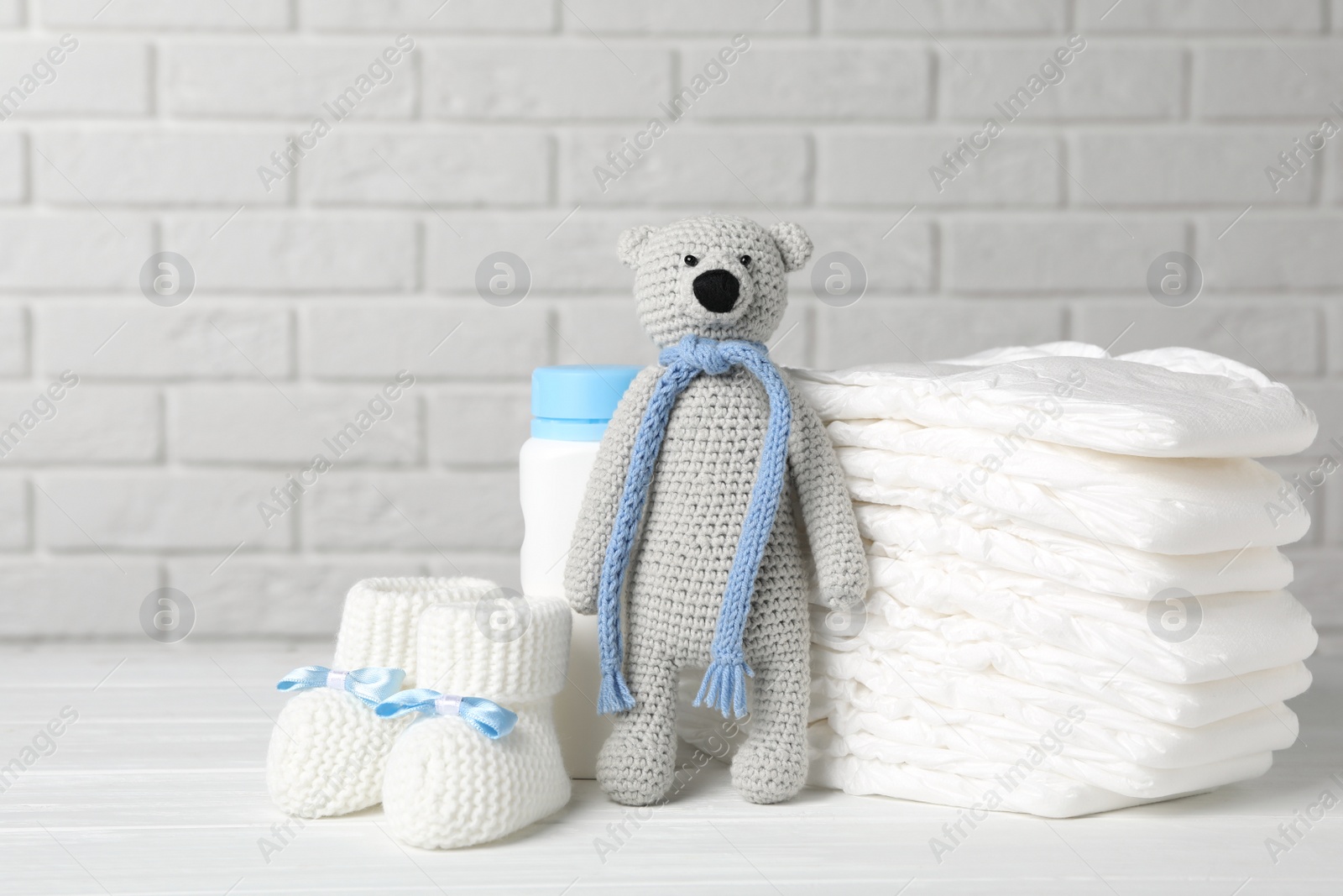 Photo of Baby diapers, toy bear, booties and bottle on wooden table against white brick wall