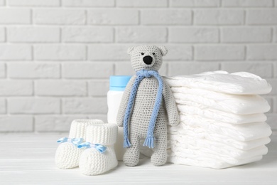 Baby diapers, toy bear, booties and bottle on wooden table against white brick wall