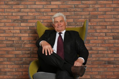 Happy mature businessman sitting in armchair near brick wall