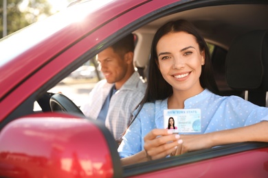 Photo of Young woman holding license while sitting in car with instructor. Driving school