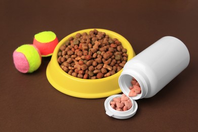 Bowl with dry pet food, bottle of vitamins and toys on brown background