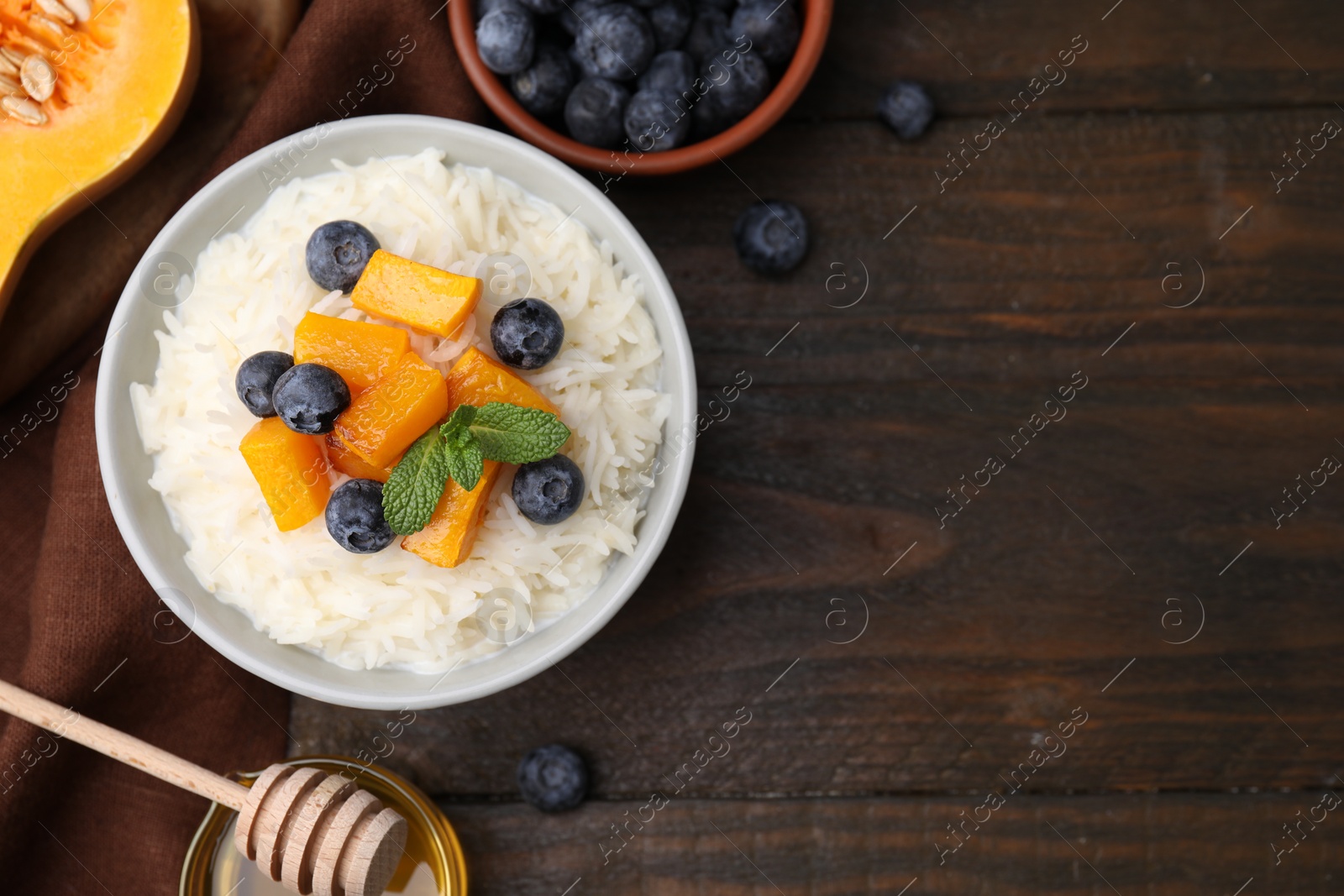 Photo of Bowl of delicious rice porridge with blueberries, pumpkin and honey on wooden table, flat lay. Space for text