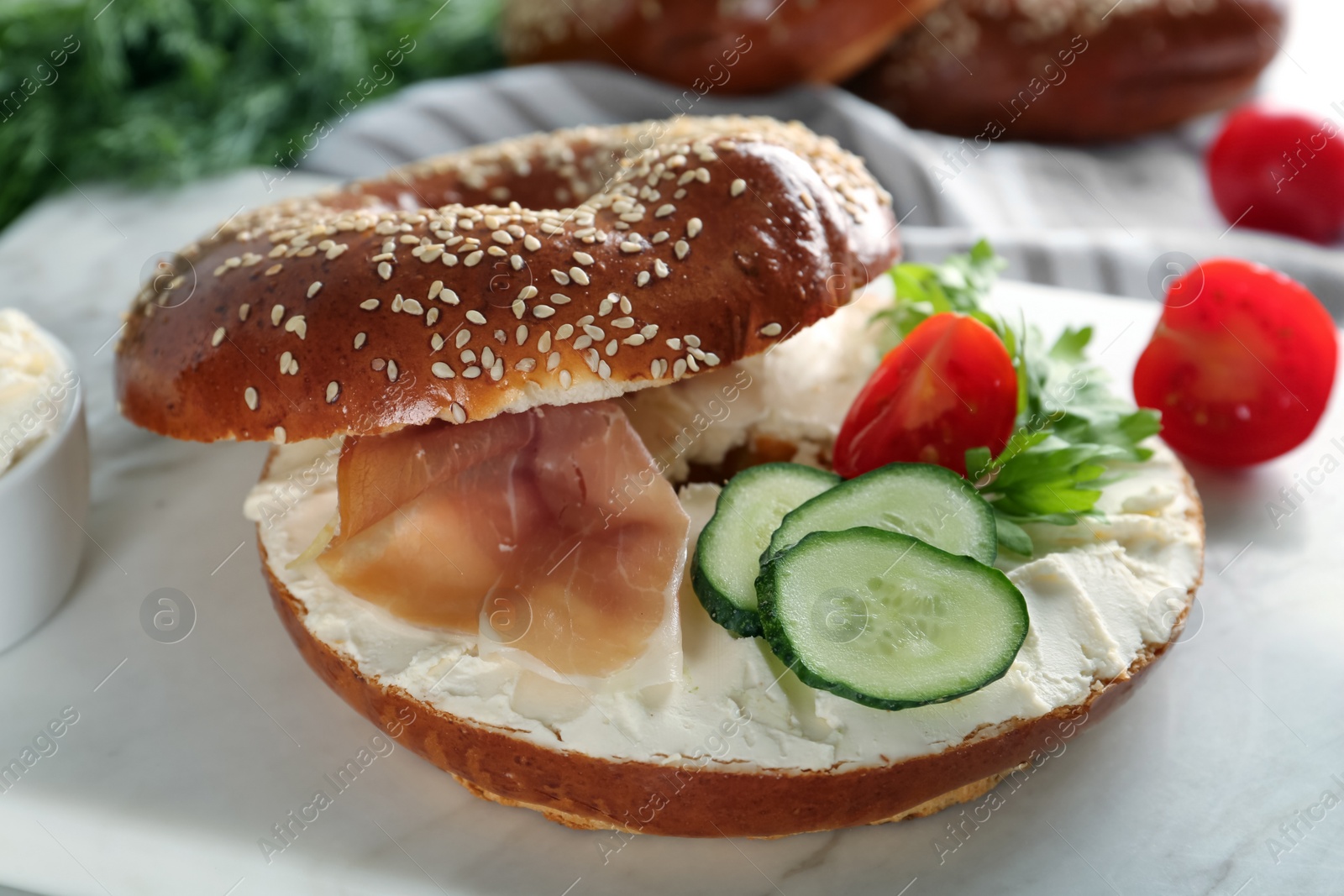 Photo of Delicious bagel with cream cheese, jamon, cucumber, tomato and parsley on white board, closeup