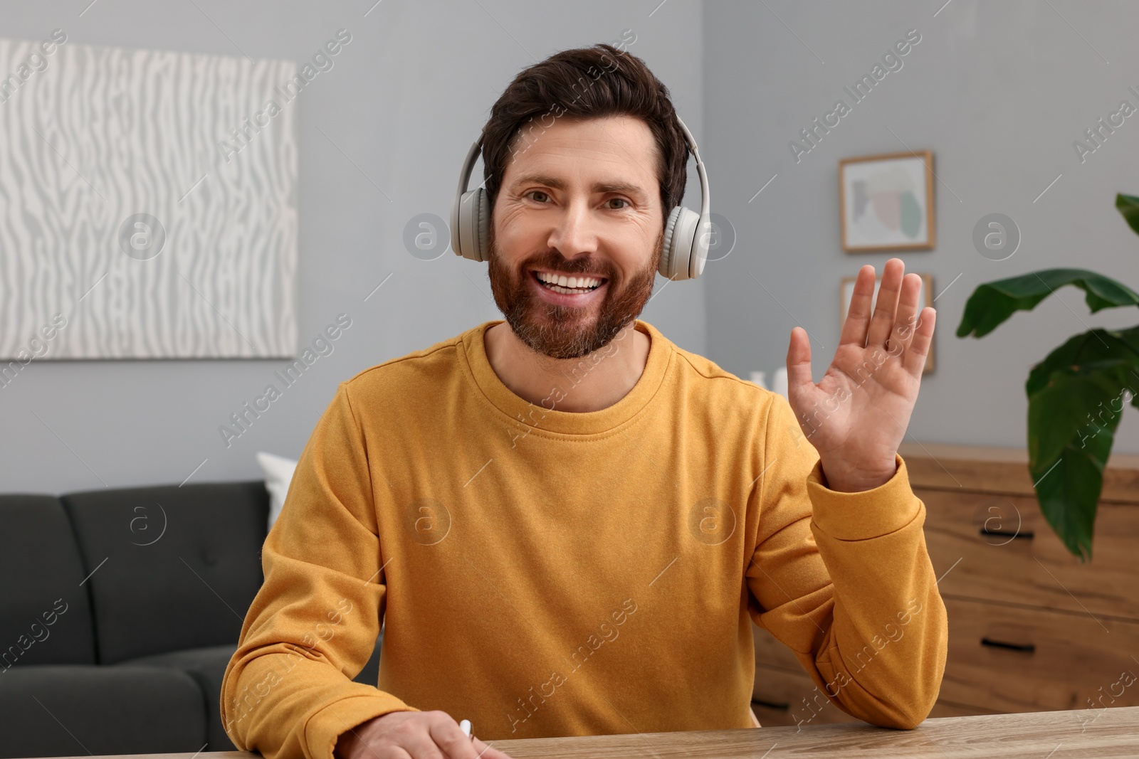 Photo of Man in headphones greeting someone at wooden table indoors