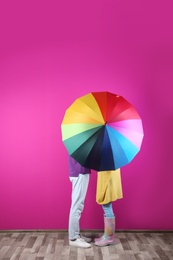 Couple hiding behind rainbow umbrella near color wall