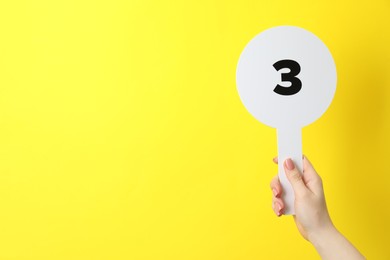 Photo of Woman holding auction paddle with number 3 on yellow background, closeup. Space for text