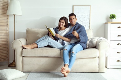 Photo of Happy couple covered with warm dark blue plaid reading book on sofa at home