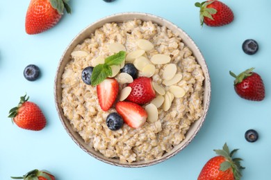 Photo of Tasty oatmeal with strawberries, blueberries and almond flakes in bowl surrounded by fresh berries on light blue background, flat lay