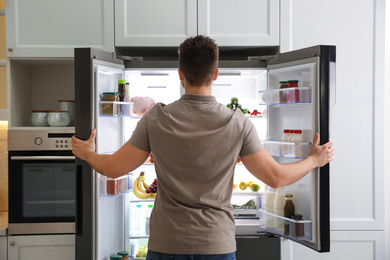 Photo of Young man opening refrigerator in kitchen, back view