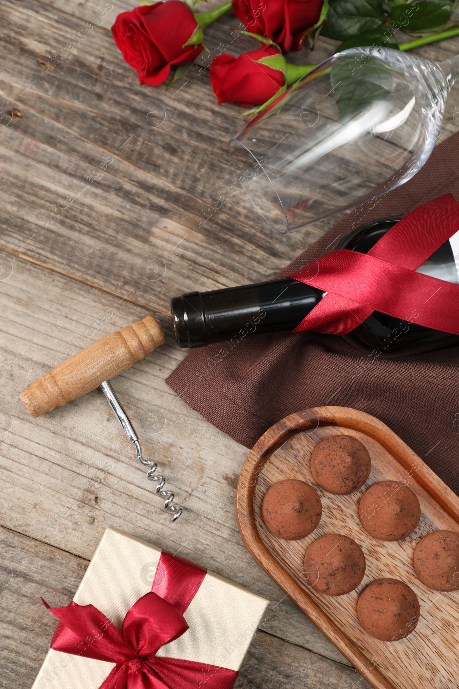 Photo of Bottle of red wine, glass, chocolate truffles, corkscrew, roses and gift box on wooden table, flat lay