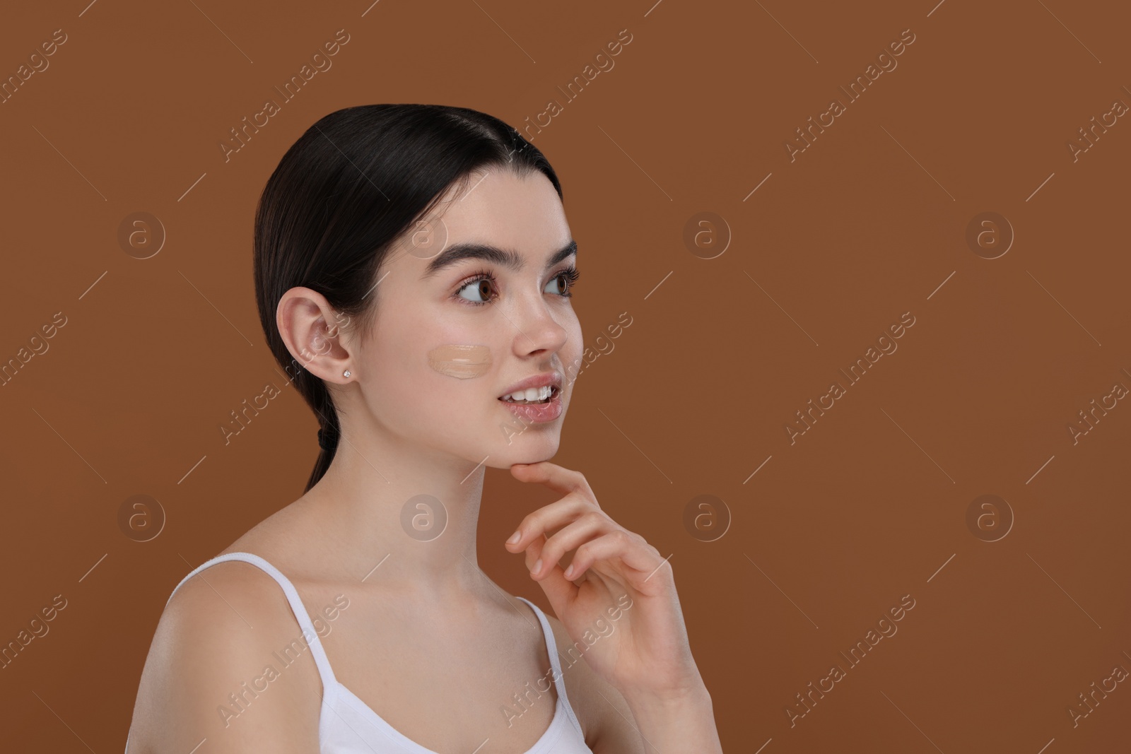 Photo of Teenage girl with swatch of foundation on face against brown background. Space for text