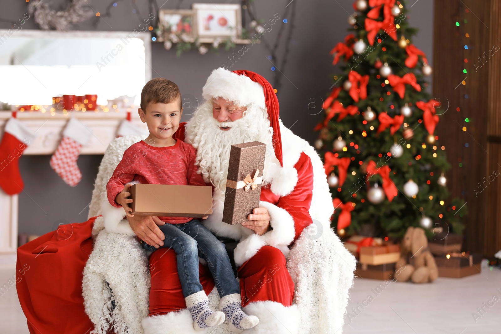 Photo of Little boy with gift box sitting on authentic Santa Claus' lap indoors