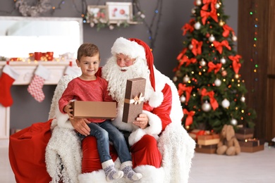 Little boy with gift box sitting on authentic Santa Claus' lap indoors