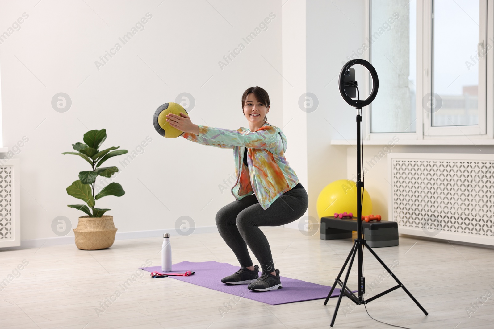 Photo of Happy sports blogger training with medicine ball while streaming online fitness lesson with smartphone at home