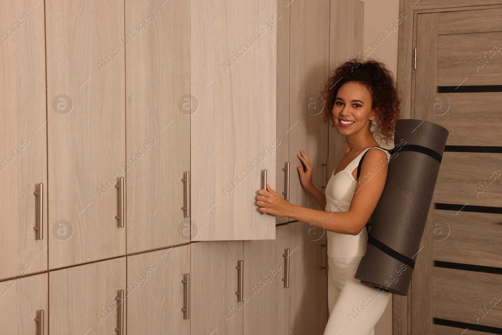 Photo of Beautiful young African American woman with yoga mat near open locker in changing room