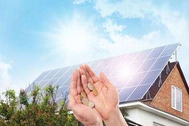 Image of Man holding coins against house with installed solar panels. Renewable energy and money saving