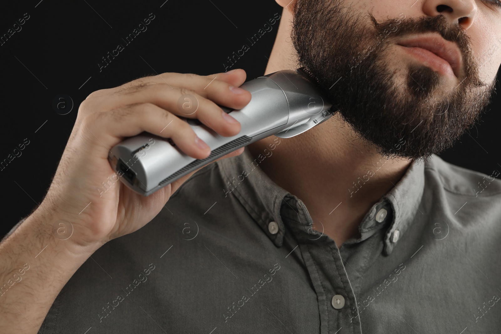 Photo of Handsome young man trimming beard on black background, closeup