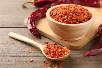 Photo of Aromatic spice. Red chili pepper flakes in bowl, spoon and pods on wooden table, closeup