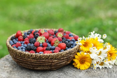 Photo of Wicker bowl with different fresh ripe berries and beautiful flowers on wooden surface outdoors