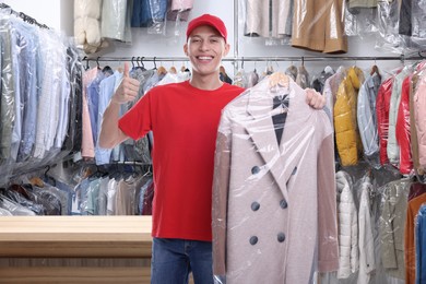 Happy courier holding coat in plastic bag and showing thumbs up in dry-cleaning